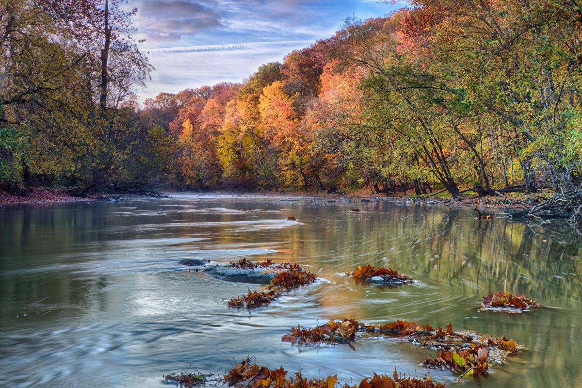 A calm river bordered by autumn trees with rocky banks, reflecting the sky and clouds above, representing the natural scenery of the Wabash River in Indiana